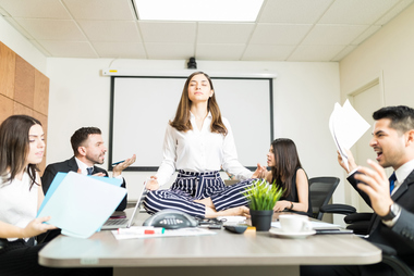 Woman meditating on office desk surrounded by colleagues