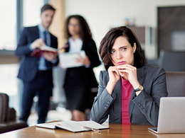 Sad female employee at desk with two colleagues looking at her from behind