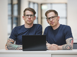 Two men who look identical sitting in front of laptop